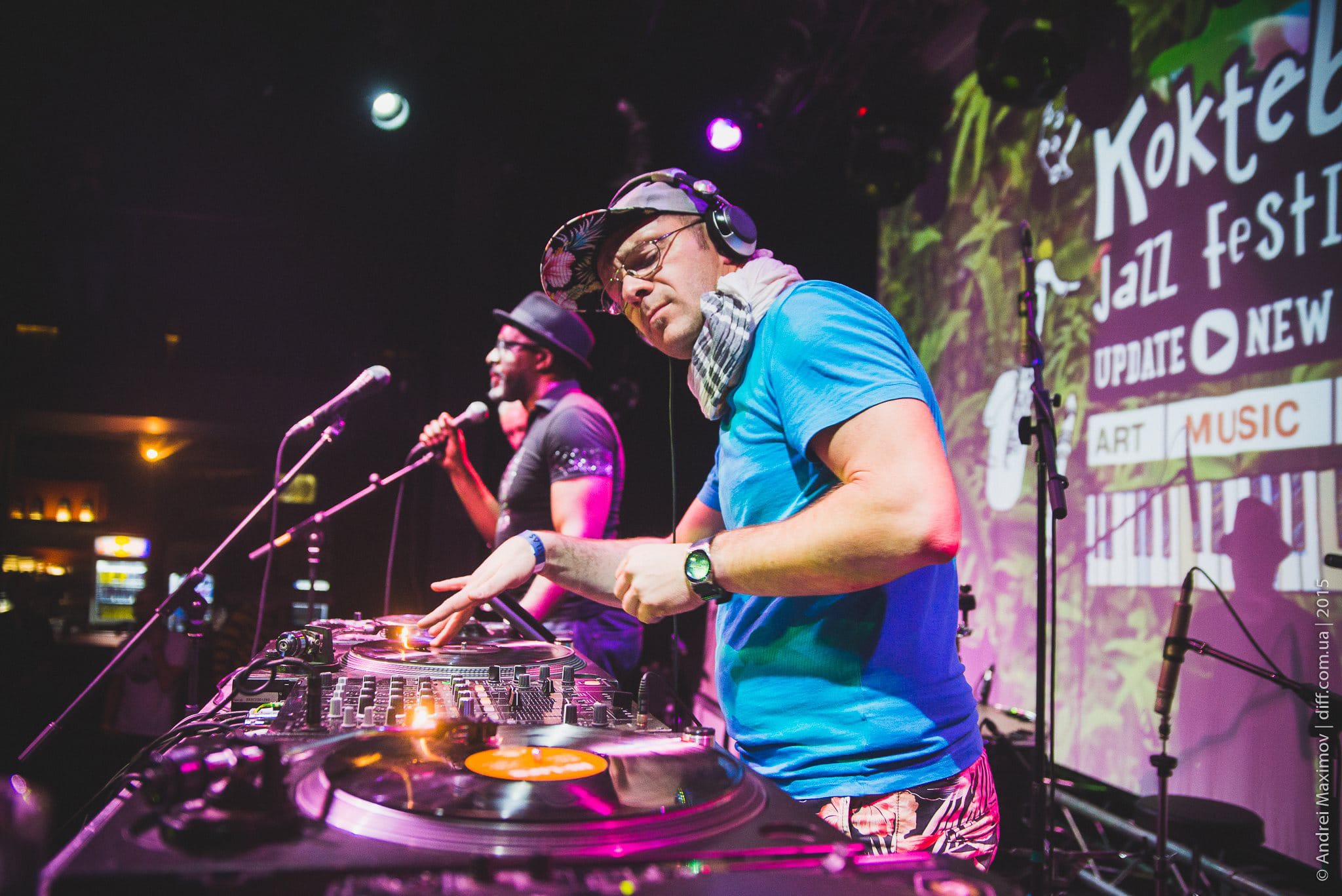DJ performing at a festival, behind decks, with a crowd in the background.